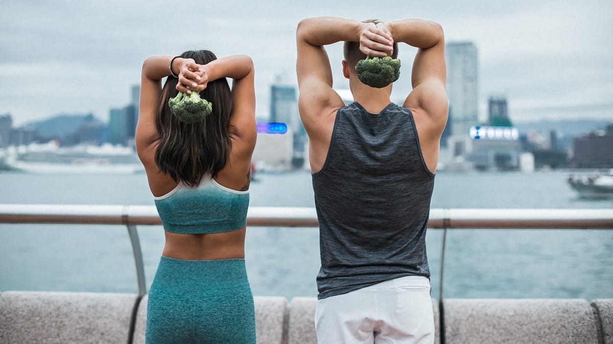Two people in athletic wear stretch outdoors by the water, each holding a piece of broccoli behind their head.