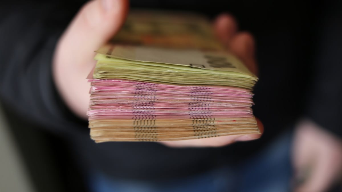 A person holds a thick stack of assorted currency notes, seen from the side, with denominations and colors visible.