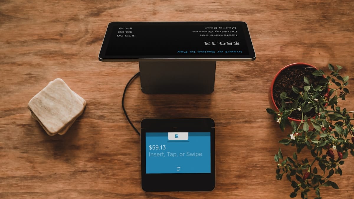 A point-of-sale terminal with a tablet and card reader on a wooden table next to coasters and a potted plant.