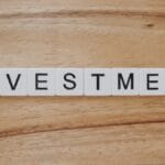 White letter tiles on a wooden surface spell out the word INVESTMENT in black capital letters.