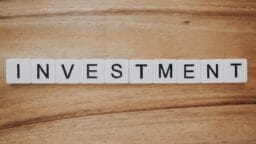 White letter tiles on a wooden surface spell out the word INVESTMENT in black capital letters.