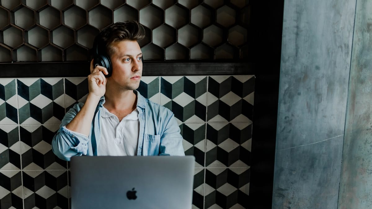 A person wearing headphones sits in front of a laptop, looking to the side, with a geometric-patterned wall in the background.