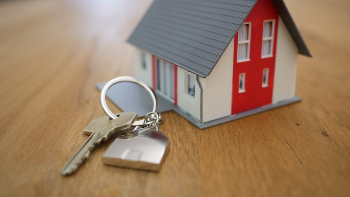 A house key with a house-shaped keychain is placed next to a small model of a house on a wooden surface.