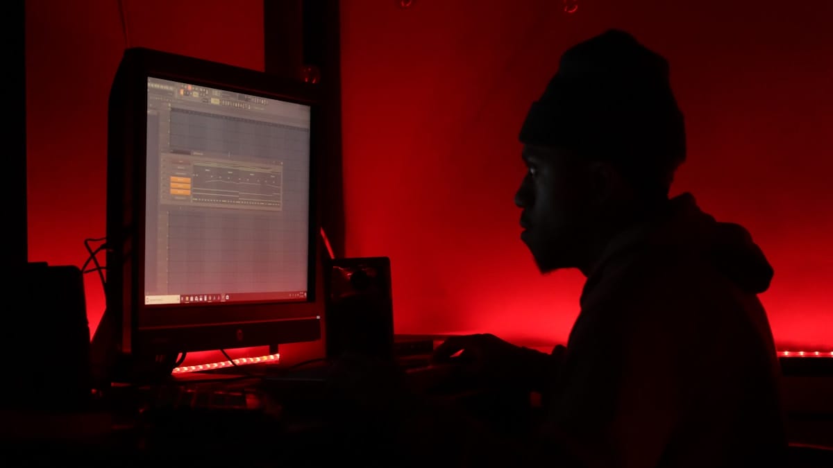 Person works at a desktop computer with music production software open, illuminated by red lighting in a dark room.