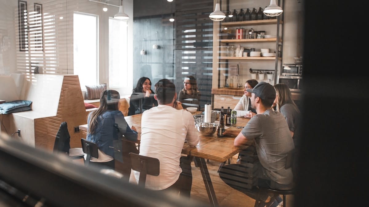 Seven people sit around a wooden table in a modern office meeting room, engaged in discussion.