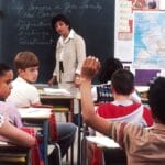 A teacher stands at the chalkboard as a student raises his hand in a classroom filled with students and colorful wall displays.