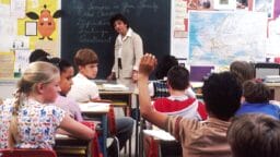 A teacher stands at the chalkboard as a student raises his hand in a classroom filled with students and colorful wall displays.