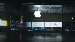 Glass-fronted Apple Store at night, with illuminated Apple logo and product displays visible inside the store.