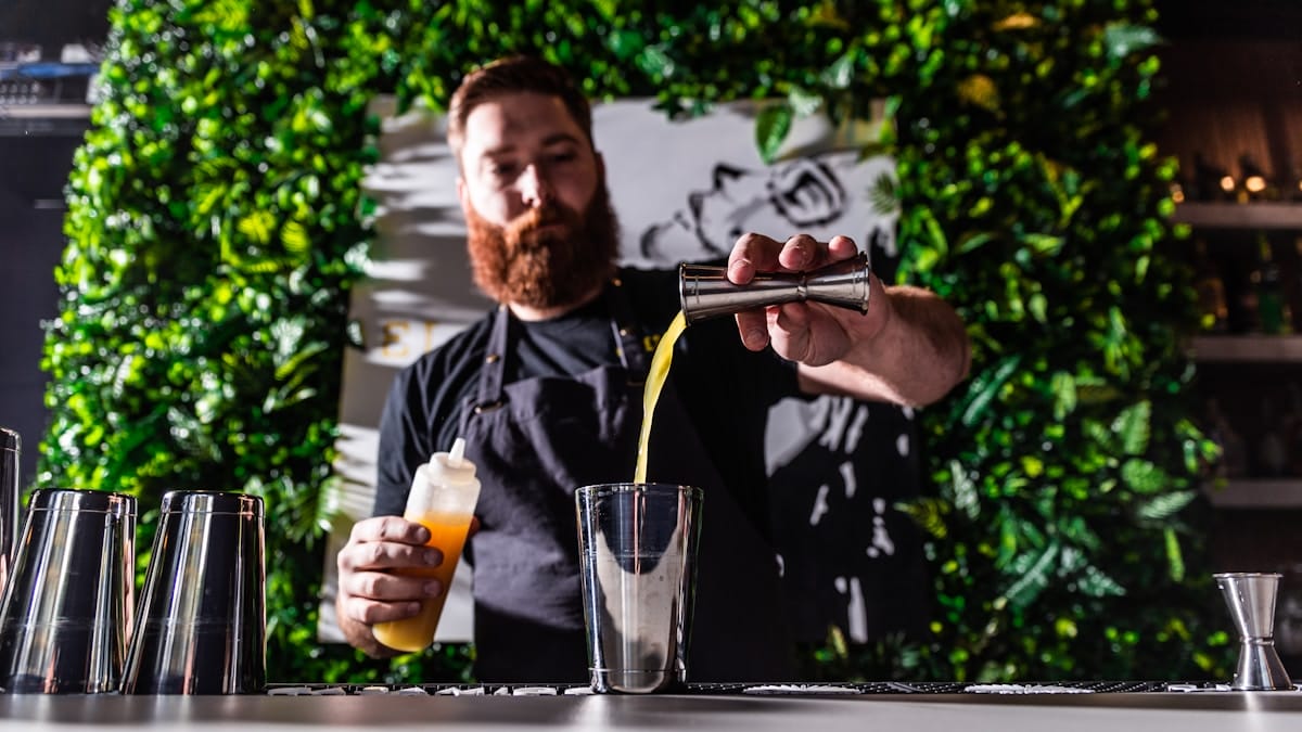 A bartender pours a yellow liquid into a shaker at a bar with a leafy green wall in the background.