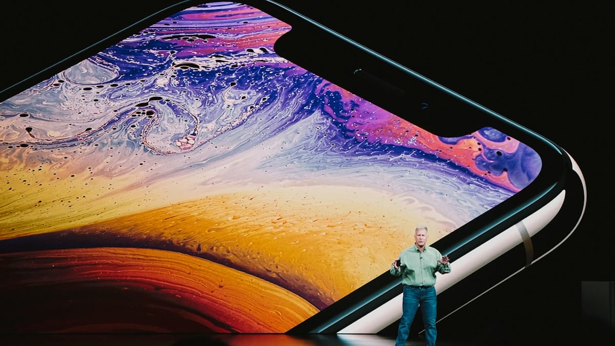 A person stands on stage in front of a large screen displaying a close-up of a smartphone with a colorful display.