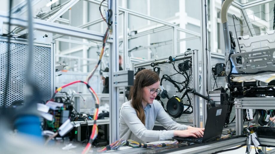 A person works on a laptop at a workstation surrounded by electronic components and machinery in an industrial lab setting.