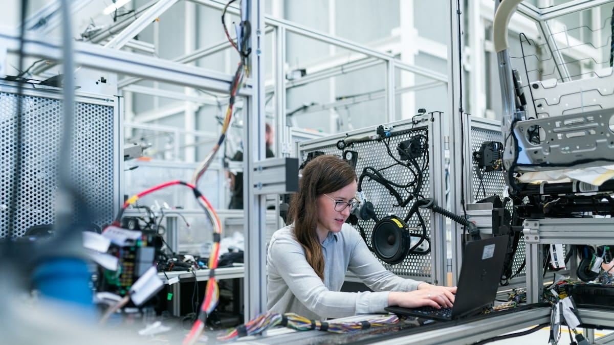 A person works on a laptop at a workstation surrounded by electronic components and machinery in an industrial lab setting.