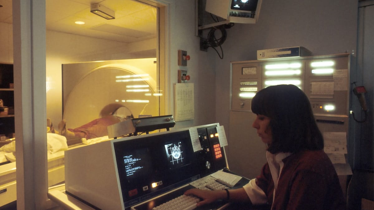 A technician operates an MRI machine from a control room while a patient lies inside the scanner.