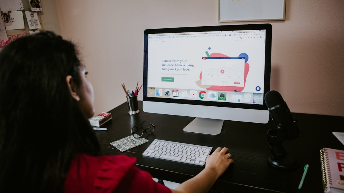 Person using a desktop computer at a desk with a microphone, glasses, and office supplies in a home office setting.