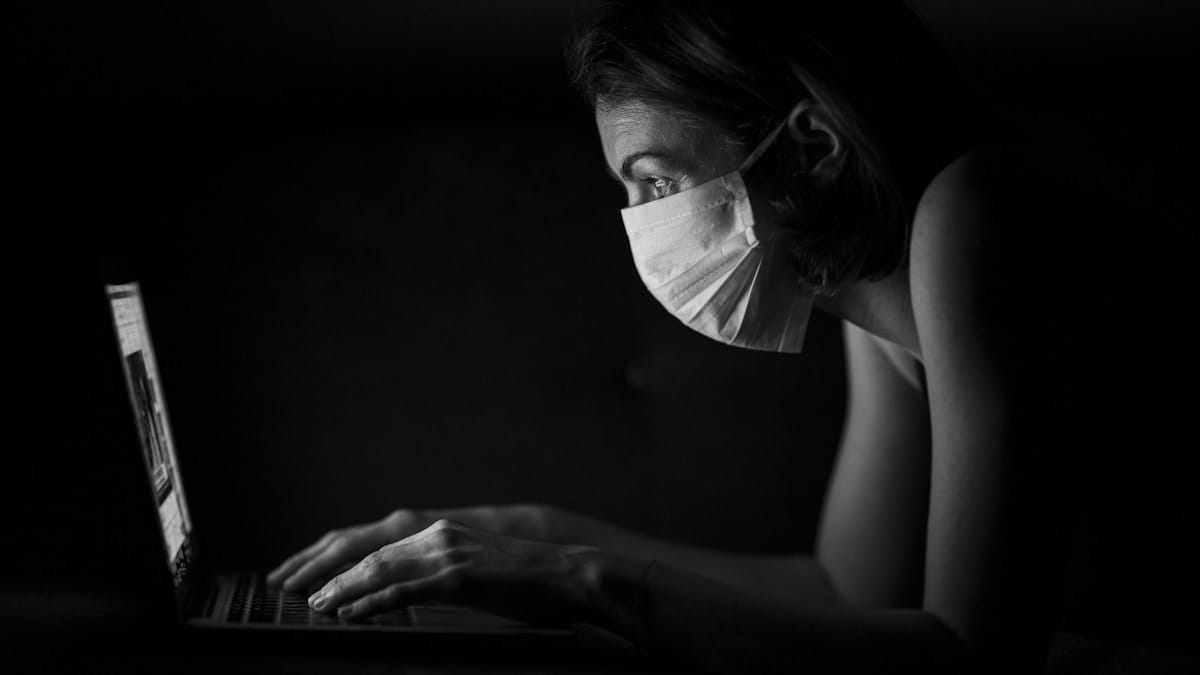 A person wearing a face mask uses a laptop in a dark room, illuminated by the screens light.