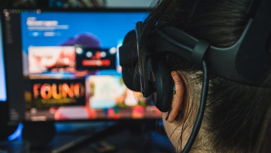 Person wearing a VR headset looks at a computer monitor displaying a selection of digital content or games.