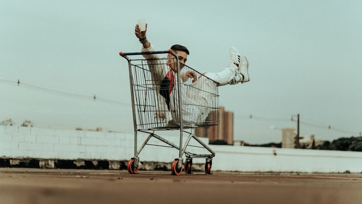 Person sitting in a shopping cart with legs extended, holding a drink, outdoors on a rooftop.