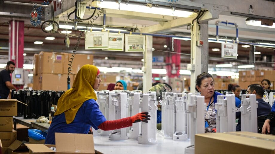 Factory workers assemble white appliances at a workbench, with boxes and machinery visible in the background.