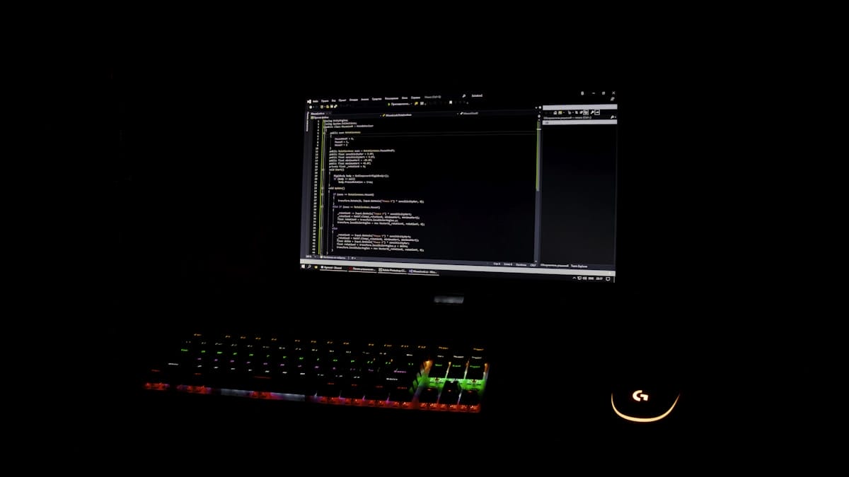 A computer screen displays code in a dark room, with a backlit keyboard and a lit-up mouse visible in front of the monitor.