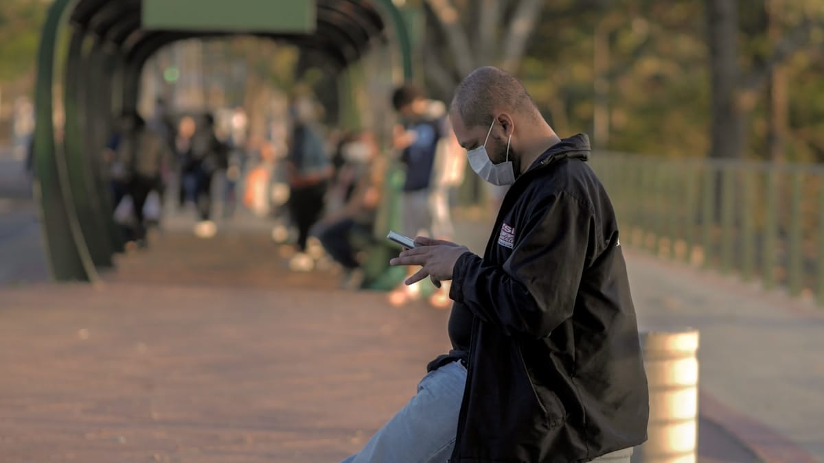 A man wearing a mask stands outdoors, looking at his phone, with people in the background on a sunlit walkway.