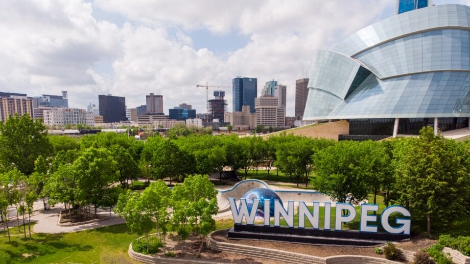 Winnipeg city skyline with modern buildings, green trees, and a large “WINNIPEG” sign in the foreground on a partly cloudy day.