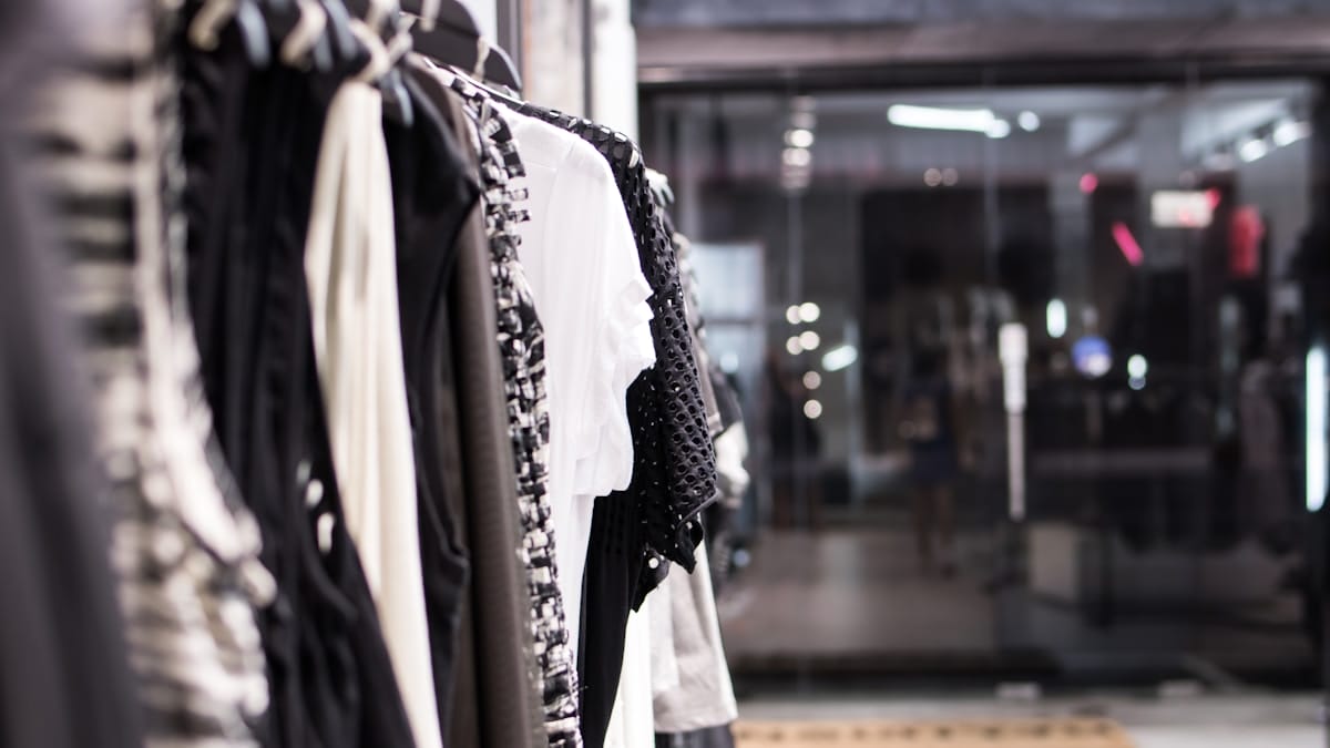 Clothes in neutral colors hang on a rack inside a store with glass doors in the background.