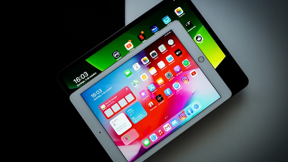 Two tablet devices overlap on a desk, both displaying colorful app icons on their screens.