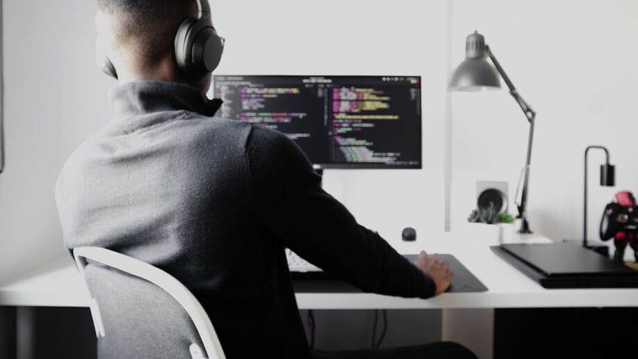 Person wearing headphones sits at a desk, coding on a computer with a dark screen in a modern, well-lit workspace.