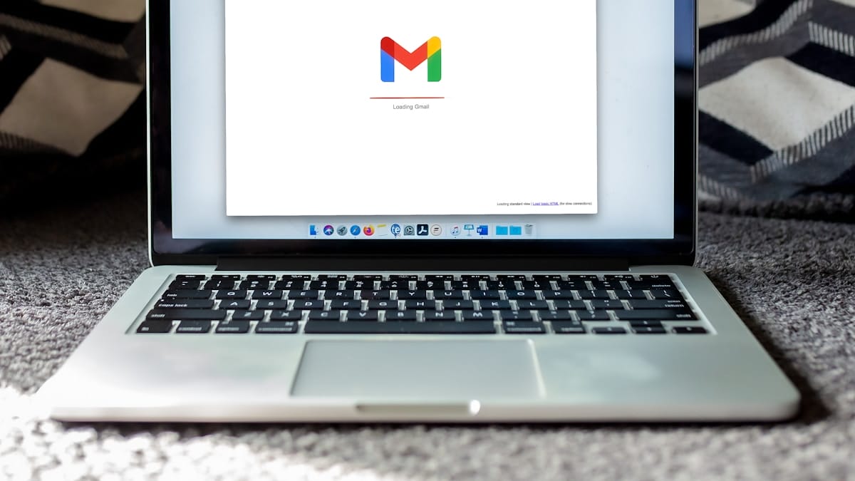 A laptop on a carpet displays the Gmail loading screen, showing the Gmail logo and the text Loading Gmail.