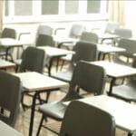 A classroom with rows of empty gray chairs and white desks, with windows in the background letting in light.