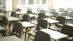 A classroom with rows of empty gray chairs and white desks, with windows in the background letting in light.