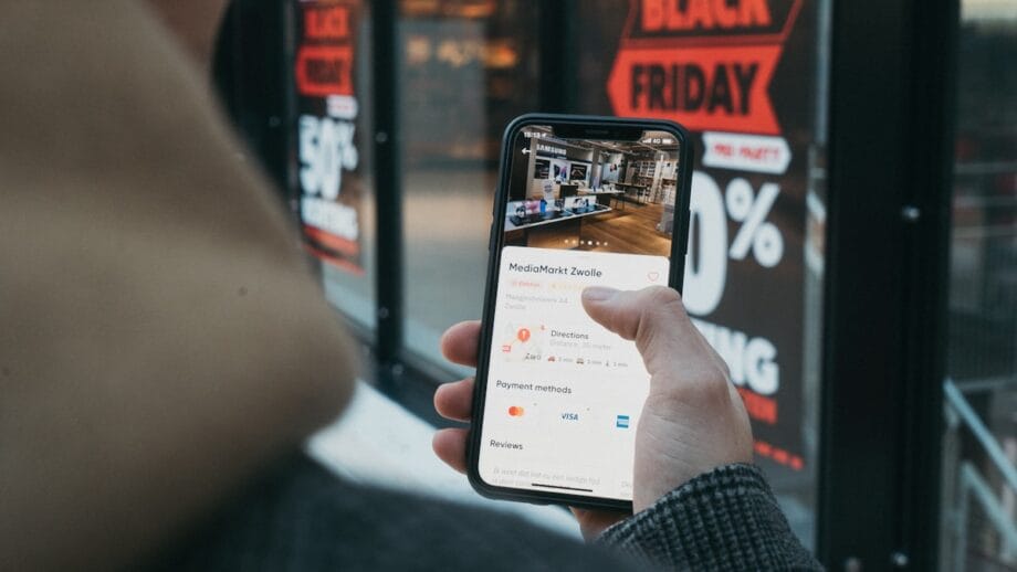 Person holding a smartphone displaying a store listing, with Black Friday sale signs visible in the background.