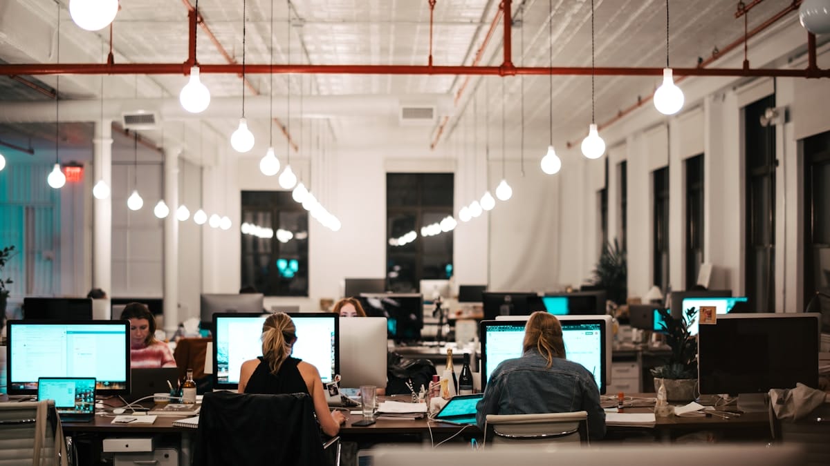 Large, modern office with people working at desks on computers under bright hanging lights in the evening.
