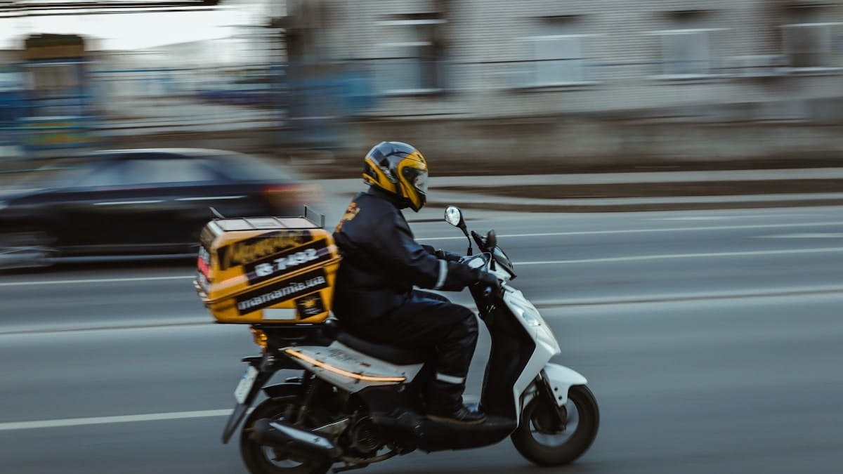 A delivery rider wearing a helmet and dark clothing rides a scooter quickly down a city street, with a blurred car in the background.