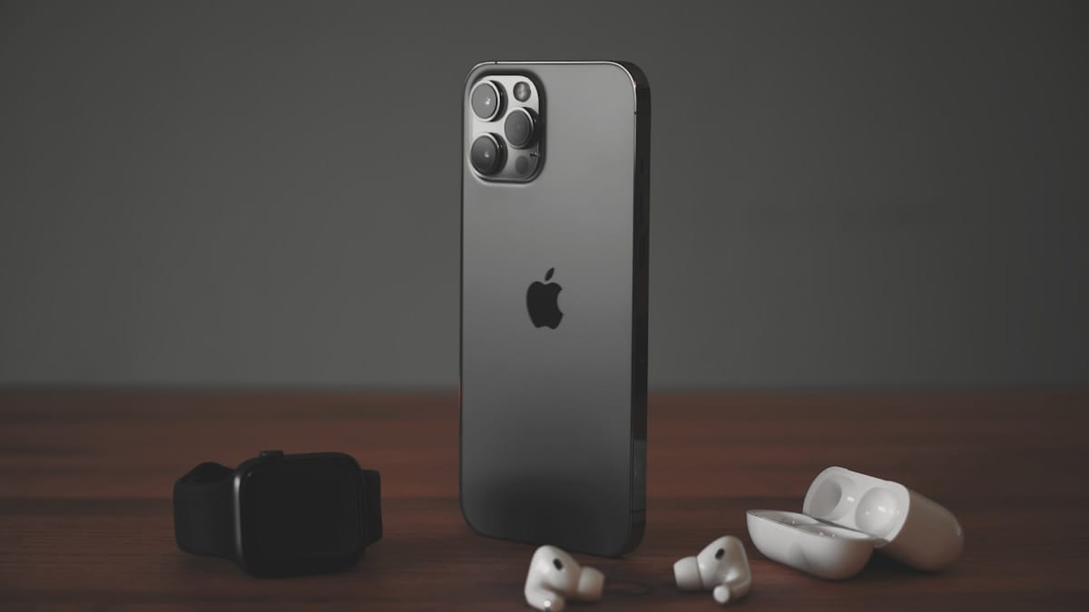A black iPhone, smartwatch, and wireless earbuds with case are placed on a wooden surface against a dark background.