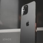 A black iPhone is standing upright in front of a Sonos speaker, showing the Apple logo and camera lenses on the back.