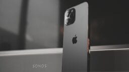 A black iPhone is standing upright in front of a Sonos speaker, showing the Apple logo and camera lenses on the back.