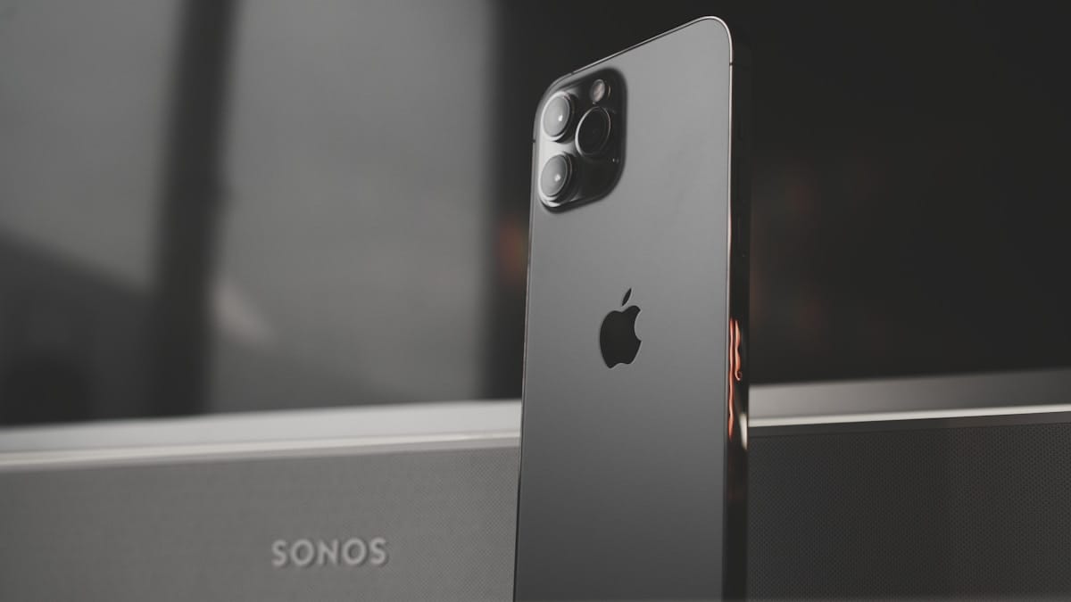 A black iPhone is standing upright in front of a Sonos speaker, showing the Apple logo and camera lenses on the back.