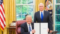 Two men in suits pose in the Oval Office; one sits at a desk holding up a signed document, while the other stands beside him.