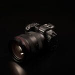 A Canon EOS mirrorless camera with a large lens is placed on a black surface against a dark background.