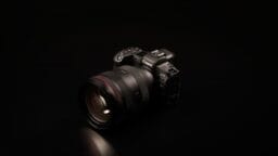 A Canon EOS mirrorless camera with a large lens is placed on a black surface against a dark background.