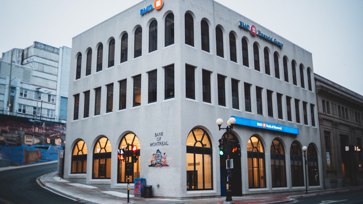 A three-story Bank of Montreal building sits at a street corner with arched windows and lit signs near a traffic light.