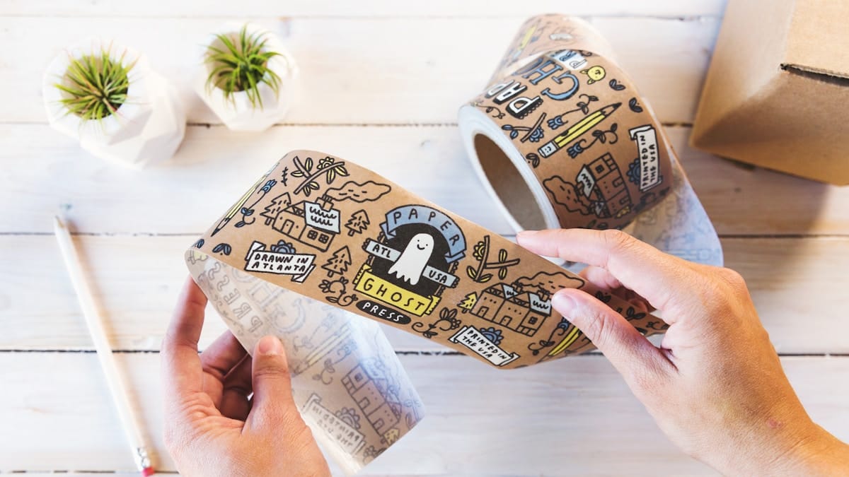 Hands unrolling illustrated brown tape with various doodles and text, next to two potted succulents and a pencil on a white table.