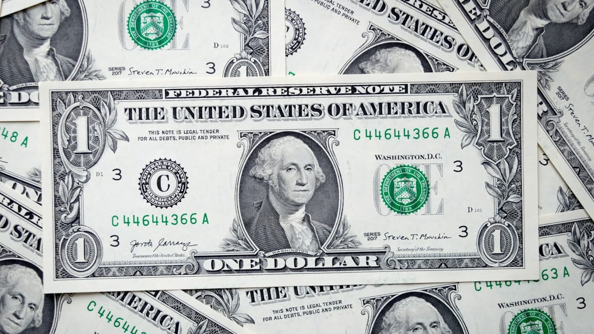 A close-up of a one-dollar bill featuring George Washington, placed on top of several other overlapping dollar bills.