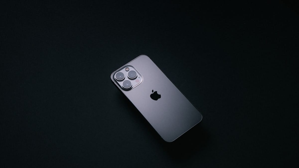 A silver iPhone with three rear cameras is placed on a dark, minimalist surface.