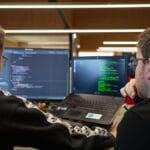 Two people sit at a desk looking at computer screens displaying code, collaborating on a programming task.
