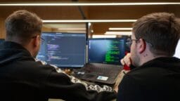 Two people sit at a desk looking at computer screens displaying code, collaborating on a programming task.