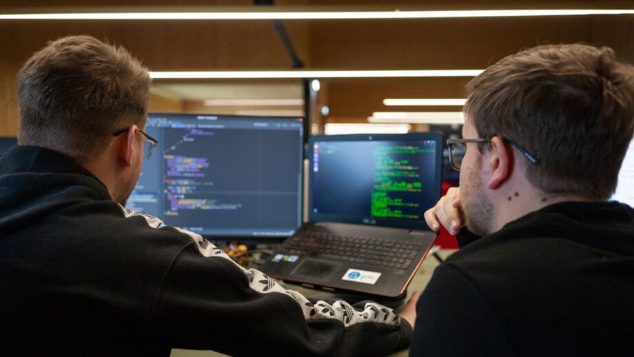 Two people sit at a desk looking at computer screens displaying code, collaborating on a programming task.
