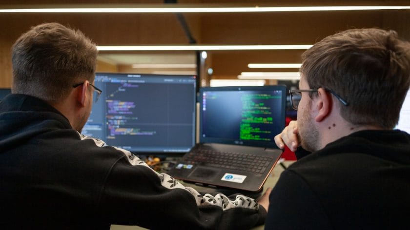 Two people sit at a desk looking at computer screens displaying code, collaborating on a programming task.