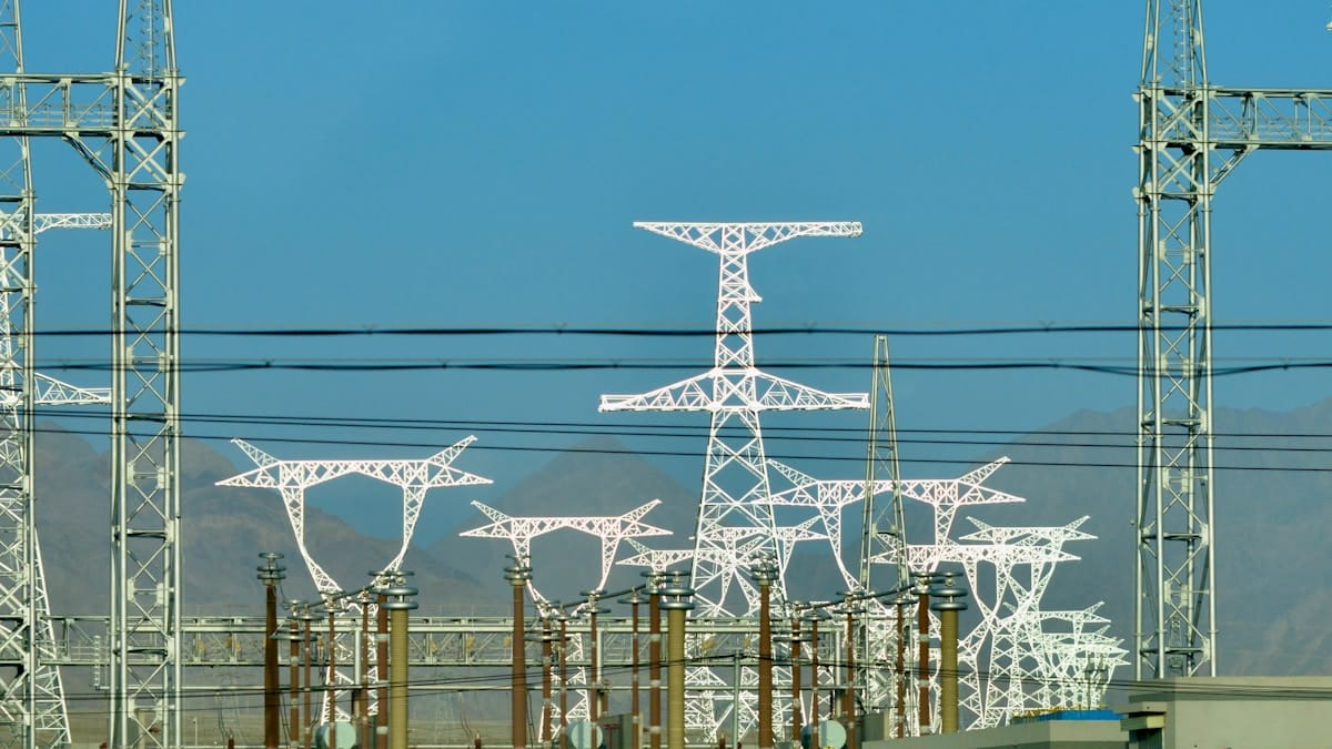 High-voltage power lines and transmission towers stand in front of mountains under a clear blue sky.